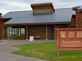 A large, brown, wood sign stands in a grass lawn in front of a large stone, wood, and metal building