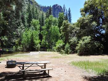 A large rock formation rises above the trees that surround a picnic table and fire pit. A rivers' edge is nearby. 