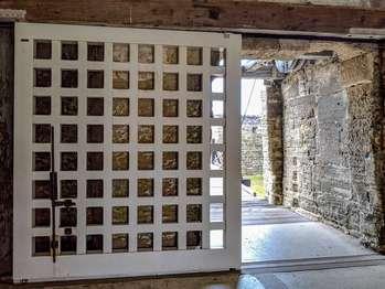 After crossing the drawbridge, the first part of the Castillo de San Marcos that people enter is the Sally Port space. This vaulted space is about 20 feet wide by 30 feet long, with an arched ceiling, about 17 feet high. The thick walls are made of blocks of shell stone. Shells are visible within the stone, but please do not touch the walls. On the upper parts of the wall and ceiling, the blocks and mortar are mostly covered with a smoother white plaster. The floor surface is smooth concrete, but slightly uneven. The description as you enter the room, going from left to right: The entrance to the Museum Store is at 9 o’clock. A wooden door frame is recessed into the wall 12 inches. The white door is built of thick wooden planks, with rounded dark metal bolts sticking out in a grid pattern. Directly across from the entrance at 12 o’clock is a 10 by 10 foot opening to the courtyard of the Castillo. On the right side of the opening is a four-foot-tall donation box with a cement base and a Plexiglas case with a hole on top. To the right of the courtyard opening, at 18 inches high, is a 15 inch deep ledge that runs the length of the wall. At 2 o’clock there is an information desk. The information desk is “Lâ€, shaped with the base of the “Lâ€, facing you. The first part of the desk is about 3 feet wide. On the left, the upright part of the “Lâ€, continues straight ahead for about another four feet. The desk has a Plexiglas top with maps and information underneath. At 3 o’clock there are two benches along the wall, side by side. They are about 3 feet long with slatted seats and backs. At 5 o’clock is a thick arched opening, about four feet wide, an accessible ramp leads you into the Spanish Guard Room. At 6 o’clock, when the Castillo is open, a large sliding wooden gate, called a portcullis is positioned to the right. It slides on rollers at the top in an overhead wooden beam and rollers on the floor. The white gate is a thick wooden frame grid, that creates a lattice with square openings about 5 by 5 inches. Hanging in the center of the door, at a 6 foot height is a 20 inch wide by 30 inch tall painting of the Castillo’s drawbridge being lifted above the moat. A dark metal rod and locking mechanism is mounted to the door just below and to the left of the painting.