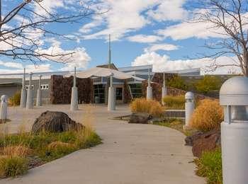 A concrete sidewalk winds through multi-colored plants and rocks to a large grey and white building