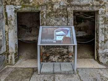 On the left side of the panel, white text against a dark brown background in English and Spanish reads:  Tidal Toilet. This colonial restroom had a twice-daily, natural flushing system. Incoming tides filled the pit with seawater, and outgoing tides flushed the waste out into the bay.  End of Text  Below, an illustration of a horizontal stone-lined channel, or sewer, runs from the left side of the panel to the right where it connects to a plaster-lined pit. Grey, rectangular stones and dark brown earth surround the pit. Half-way up from the pitâ€™s bottom, is a wooden plank with 3 holes cut into it for seats.   The panel sits just inside the stone opening to the fort's former toilet room. Large blocks of light grey, brown, and tan stone frame this opening. Beyond the panel, are views of the room's mottled grey plaster walls and dirt-covered floor.  [End of message]