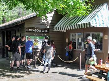 People stand in line beneath a sign that reads, 'Meadow Grill,' on a deck with picnic tables.