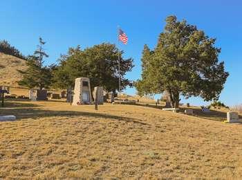 Cemetery on a grass and small tree covered hillside with numerous headstones and a monument
