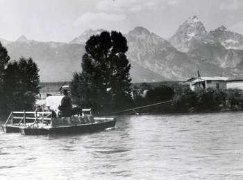 Historic image of Menors ferry carrying a covered wagon crossing the Snake River toward the west. The cabins and the Teton Range lie beyond.