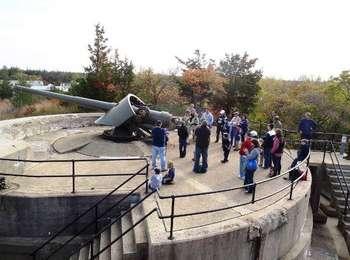 A crowd of autumn visitors inspect the barbette gun atop the gun emplacement, overlooking the dunes and native vegetation.