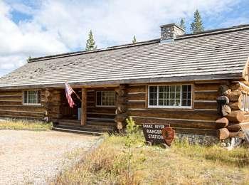 A gravel lot leads to rustic log cabin with an entry way, US flag, and wood shingles