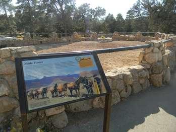 A tilted interpretive exhibit board mounted in a metal frame supported by two metal posts stands next to a low, circular rock wall. Large black letters at the top of the board read