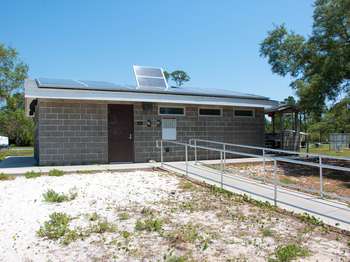 A grey stone building with a brown door. and small windows. Solar panels are attached to the grey roof. A cement sidewalk with metal railings in the foreground. Green trees and blue sky in the background.