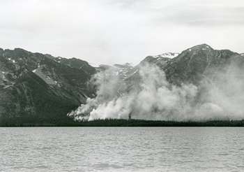 A black and white photo shows a fire burning on a hillslope across a lake.