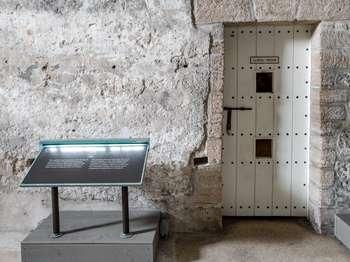 The carcel or prison is visible through small windows in a locked door on the east wall of the Spanish Guard Room. The 6 foot tall, wood paneled door is white, with black metal bolts, and a sliding metal locking bar. In the center, near the top and bottom of the door are two openings about 5 inches wide by 8 inches tall. The upper opening, at a 5 foot height, has a metal screen. The lower opening, at a 3 foot height, is unscreened. A metal plaque on the door reads,   Carcel-Prison  The view of the carcel, as seen through the door openings, shows a room 10 feet wide, 12 feet deep, and 10 feet tall, with an arched ceiling, and an uneven dirt floor. The shell stone block walls are partially covered with smooth plaster. Carvings are seen on both the plaster and the stone blocks. The room has no windows but is lit for visitor viewing.   The carcel door has an exhibit on either side, both in English and Spanish. The exhibit on the left is titled,  Prison Carcel: The text reads,   This small cell acted as the town jail for the Spanish and as a military jail under British occupation. During the American Revolution, South Carolina statesman and patriot Christopher Gadsden was held in solitary confinement here for 42 weeks.  The exhibit on the right is titled,  Juan Oâ€™Donovan. ca. 1760 to ca. 1820.  A portrait shows a lieutenant in the Spanish Hibernia Irish Regiment, described in detail later. Text explains that this man fell in love with a governorâ€™s daughter and eloped. Because the governor disapproved of the match, Oâ€™Donovan was arrested and sent to Havana, Cuba. After two years, the governor relented, and Oâ€™Donovan returned to be re-joined with his bride.   In the portrait, Oâ€™Donovan seems to be bowing slightly. He has rosy cheeks, a high forehead, brown eyes, and a rounded nose. He wears a white wig that has a circular curl at ear level. He is wearing a red jacket, vest, and breeches that reach just below the knee. His shirt has a high white collar that covers most of his neck. Two inches of ruffled white lace show above the vest and wrists. A gold and black sash is worn across his jacket, from his right shoulder to his left hip. A sword, with a hilt with a gold tassel, is worn at his left hip with the tip of the sheath protruding beyond the tail of his jacket. He wears tall white knee socks, with black shoes that have a wide gold bucket on the top. In his right hand, he holds a black hat that curves up in three corners, to make a triangular shape. His left hand rests on the sash across his chest, near his heart. 