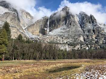 A small dusting of snow rests around Upper Yosemite Fall, which is a thin strand of water falling. Clouds hang low along the granite walls.