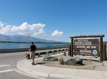 Visitor standing near Jackson Lake Dam sign with Jackson Lake and northern Teton Range beyond.
