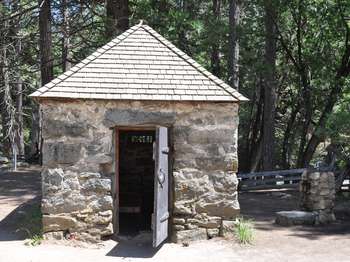 An old cot can be seen through the open door of a small stone structure with a pointed roof.