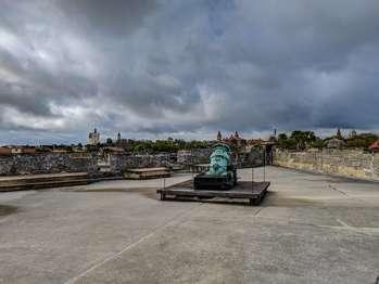 When viewed from above, as from a bird’s eye, the Castillo is square in shape, with four diamond-shaped bastions extending from each corner. These diamond shaped bastions are connected by the gundeck. The San Pedro Bastion is located on the northwest corner of the Castillo. The view from this bastion includes the moat, the grassy area surrounding the Castillo, and the parking lot. The historic city and the original stone pillars that held the city gate can also be seen from this location. The bastion has low walls, about 5 feet tall. Each side of the diamond-shaped structure is unique. Facing the bastion from the gundeck, going from left to right: Along the east facing wall there are two cutouts in the wall, with a two-step platform between, each about 2 ½ feet tall and extending out from the wall about 4 feet. Between the platforms, the upper walls have cutouts about three feet wide by three feet high, stopping at the platform level. On the south facing wall there are three cutouts with two-step platforms between them. A cylindrical shaped sentry box with a domed top is at the southwest corner. It is closed to the public by a wooden barricade. The west facing wall no two-step platforms along this side, with one cutout at the north end of this wall. The north facing wall has one four foot wide cut out with no two-step platforms. In the center of the bastion is a 9 foot square, low wooden platform. A rope stretches between metal posts at each corner. In the center of the platform is another wooden platform with a 15-inch bronze cannon, called a mortar. The bronze has weathered into a pale green patina color. The tube-shaped mortar barrel is tilted up into the air at about a 45-degree angle. Its base is connected, crossways, to a thick bronze rod. This rod is secured to the wooden platform by thick metal strips with rounded metal knobs. The front of the mortar platform has a raised wooded lip with metal strips securing it on each end. The mortar barrel has ornate bronze sculptures. Near the base is a bearded face and the date 1724. At the center of the barrel is a coat of arms. At the top, are two curved handles in a dolphin shape.