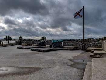 If viewed from a bird’s eye, overhead, the Castillo is square in shape, with four diamond-shaped structures extending from each corner. The diamond shaped structures, called bastions, are connected by a path called a gundeck. The San Agustin Bastion is located on the southeast corner of the Castillo. The view from the San Agustin Bastion is of the St. Augustine Bay (or Bayfront). The entrance, fee booth, the sea wall, and the parking lot can be seen from this location. The east facing bastion walls are about one to two feet tall and the south facing walls are about 5 feet tall. Each side of the diamond-shaped structure is unique. Facing the bastion from the gundeck, going from left to right: At the southeast corner, is a cylindrically shaped structure with a domed top. You can walk to the structure through a 10-foot corridor. There is a low doorway, approximately 5 ½ feet tall. You may need to duck down. The interior is 8 feet high with three small square windows. The south wall has a two-step stone platform that runs along the wall, it turns the right corner and continues to the corner of the gundeck. There are also wooden platforms, about 7 to 8 inches, that extend out from the wall about 18 feet. Two ornate bronze cannons sit on top of wooden carriages which stand about 3 feet tall and have four wooden wheels wrapped with iron straps. They face out to the historic city though cutouts in the wall. To the right of those cannons is a pole flying the Spanish colonial flag, which is white with a ragged red “xâ€, shape. The right corner of the diamond shape has two 4 foot wide cutouts in the wall before the bastion connects to the gundeck.