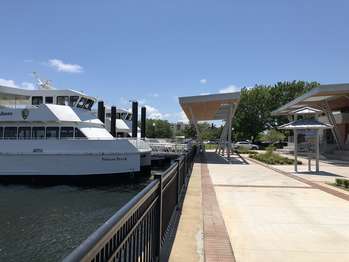 A cement walkway leads up to metal structures and an interpretive wayside. Two white tour boats are docked in the water on the left side.