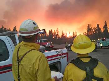 Two firefighters stand in a large parking lot watching an intense crown fire approaching.