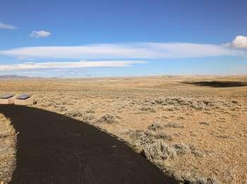 A paved path with two wayside signs is in front of a sagebrush and dried grass plain that stretches between two ridgelines