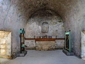 The Chapel is accessed from either the Second Spanish Room or the American Indian room. This roomâ€™s exhibits, in English and Spanish, describe religion at the Castillo. This room is described as if entering from the Second Spanish room.  The courtyard entrance has a wooden door that forms an arch and opens inward to the room. That door remains closed. Above the door is a window.   The room is about 20 feet wide and 30 feet long, with an arched ceiling about 17feet high.  In the center of the back wall, a 5 foot tall arched niche is recessed about one foot into the wall. A narrow four foot ledge runs the length of the back wall with a five foot long, 4 foot section in the center. All ledges have a wooden top and trim. The altar has 4 images of the chapel during different time periods.  The entrance to the American Indian room is on the east wall.  Standing at the Second Spanish Room entrance to the room, the description, moving from left to right, includes:  Religion at the Castillo Exhibit. An illustration of a religious service with soldiers attending. Illustrations of the chapel. Text explains that this chapel was the scene of daily worship. Religion guided all aspects of colonial life. Roman Catholic Christianity was the common faith throughout the Spanish empire.  LÃ³pez De Mendoza Grajales. 1530 â€“ 1590. Text explains, in 1565, Father LÃ³pez De Mendoza Grajales celebrated the first mass in St. Augustine that would become the United Statesâ€™ oldest parish. The portrait shows an older priest standing behind a wooden table wearing a gray robe. He is partially bald, with a tidy, thin circle of hair around the bare crown of his head. The robe is long sleeved with a simple knotted white rope at the waist. He holds an embossed black book. A skull rests next to his arm on the table.  What do the Walls Tell Us? Exhibit. A vertical glass panel protects an ornate arch inset in the wall. Below, a photo shows holes in the stone wall. The text explains that Catholics dip their fingers in Holy Water and make the sign of the cross before worship and prayer in the Castilloâ€™s chapel.  On the south wall of the Chapel is a closed, inaccessible door to the courtyard.  What do the Walls Tell Us? Exhibit. A vertical glass panel protects an ornate arch inset in the wall. Below, a photo of two men outside a wooden doorway with an arched top. The text explains that behind the glass are the remains of a plastered holy water font.