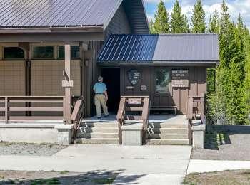 Man in turquoise shirt and tan pants walking into front door of building at the top of four steps marked Backcountry permits.  A sign on left side of building says visitor center to the left.