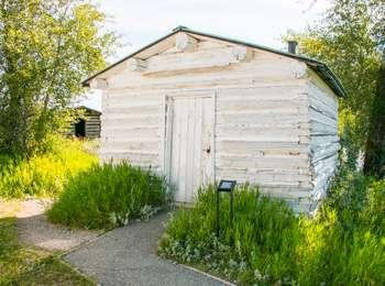 Front of a white painted log building with a door that is 12 feet square with a low pitched roof.  A small exhibit panel is to the right of the door.