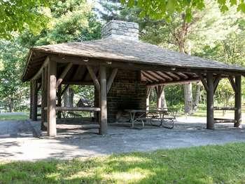 A color photograph with a wooden pavilion containing a large central stone fireplace, picnic tables, and a slanted roof.