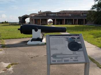 Metal framed National Park Service sign in front of a large black cannon. Brick Fort Pickens walls in the background.