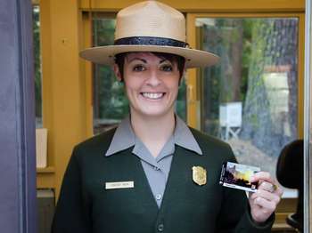 A female park rangers is holding up a Yosemite National Park Pass and smiles at the camera.