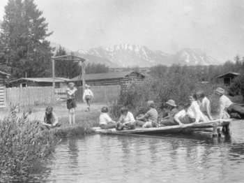 Several young people lounge on a log diving board, which extends several feet out into a swimming pool.