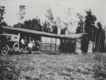 Six people stand on the porch of a log cabin in the trees, next to an antique car.