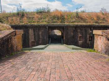 Brick pathway leads across a green wooden drawbridge to an archway opening in the side of a brick fort.