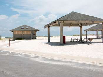 Two light brown pavilion structures with brown picnic tables underneath on the right. A light grey stone building with a brown letter