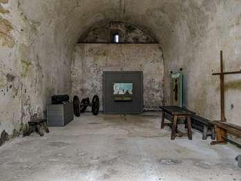 The Siege Room is accessed from the courtyard with a slight upward slope. The siege room exhibits show how the Castillo’s cannons were used to defend the fort. Most exhibits are in English and Spanish. A double wooden door opens into the Siege room. Above the door is a slight ledge. Above the ledge is a barred window with a wooden shutter that also opens inward. The room is about 20 feet wide and 30 feet long, with an arched ceiling about 17 feet high. Standing at the entrance to th room, the description, moving from left to right, includes: Soldiers Through Time Exhibit. Several modern photographs of re-enactors dressed as soldiers from the Spanish Era to the World War 1 Era. Deadly Explosion 1702 Exhibit. Part of a tactile cannon barrel sits on a cement and wood pedestal. The text explains that the 18 pound iron cannon was uncovered in the moat. In the corners of the back wall are two tactile cannons on carriages with large wheels. These are working replicas, so please do not climb on or move pieces of them. Small wooden barrels are located between the wheels of each cannon. Defending the Fort 1740 Exhibit. A 3- dimensional, miniature, diorama shows soldiers, each about 6 inches tall, peering over the northeast bastion wall preparing to fire a cannon. Text describes a battle in 1740. Francisco Menéndez. 1705 to 1770. Text explains that Menéndez, a former slave, escaped to St. Augustine in 1724. By 1738, he had become a captain in the militia and commanded the first free Black settlement in North America. An illustration shows him wearing a black triangular hat over curly jaw length hair. He has an angular face. He is wearing a long black jacket with gold closures over a long red vest and black breeches, that end just below the knee. White fabric is wrapped around his neck. Two inches of ruffled white lace shows above the vest and wrists. He wears tall white knee socks, with black shoes with a wide gold buckle on the top. His right hand rests on thin cane with a tassel at the top. He holds the hilt of a black sheathed sword in his left hand. A tactile wooden gun rack. Spanish Colonization Exhibit. An historic map of the ocean off the coast of Florida. An illustration of a man in a blue uniform with red cuffs and socks, wearing a black triangular hat, and holding a musket. Historic maps of St. Augustine. The text explains that the first colonists in St. Augustine endured hunger, privation and disease. Yet it was critical to the Spanish Empire as a base to patrol Florida’s coastline for pirates, rescuing shipwrecked survivors and salvaging wrecks.