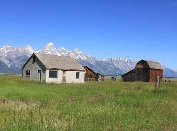 Thomas Murphy homestead with gray stucco house, out buildings, and large brown barn surrounded by grassy field. Visitors standing on the far right of the barn and the Teton Range panorama behind.