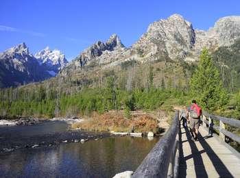 Hikers on bridge across the outlet of String Lake just past trailhead with view of the Cathedral Group and Mount St. John