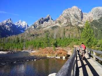 Hikers on bridge across the outlet of String Lake just past trailhead with view of the Cathedral Group and Mount St. John