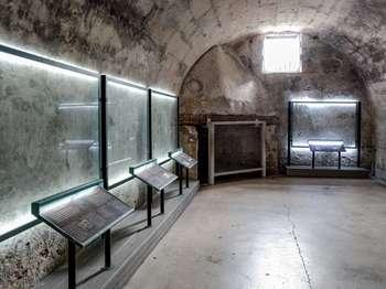 This second Spanish Guard room is accessed through the first Spanish Guard room. A white wooden door, with black bolts and a sliding metal locking bar, opens into the room. The room is about 20 feet wide by 30 feet long, with an arched ceiling about 12 feet high. The room has several illuminated glass panels that frame sections of the walls. It also has a window, fireplace, and several exhibits. All exhibits are in English and Spanish. If, after entering the room, you turn to face left, this describes the room, going from left to right: Along the left wall are three of the illuminated glass panels. In front of each is an exhibit relating to wall carvings behind the glass. A brief summary for each exhibit panel includes: A Spanish Fleet: A sketch of a sailing ship. Text explains that Spanish soldiers made the carvings of the fleets on the walls. American Flags on Foreign Ships: A sketch of a sailing ship with a striped flag mounted on the stern. Text explains that British ships like this are carved into this wall and the north wall. U.S. soldiers may have added American flags to the carvings. Etched, Burned, and Scrawled: Photos and sketches show letters of the alphabet are carved into the wall. Some are written backwards. Text explains that this wall contains 200 year old graffiti left by former soldiers. Soldiers of the 18th century were usually illiterate, so the artists may have just been learning to read and write. At the left corner of back wall is a curved block fireplace with a smooth top shaped like a half cone. The opening to the fireplace is protected by a clear plastic panel. At the top of the back wall is a 5 foot by 6 foot window, with metal bars, and shutters that open inward. Below the window is another illuminated glass panel and an exhibit, titled, British Ships: A line drawing shows a sailing ship with a large striped flag. The text explains that although we don’t know who made the ship carvings, it could have been Spanish soldiers. They would often have seen British ships in battle. An American soldier probably added the U.S. flag. On the right wall is the entrance to the carcel, or prison. Exhibits on either side of that door are audio described separately. Behind you, on the back wall, is another exhibit panel, titled, A Mysterious Note: A diagram shows several, faded, handwritten words. Text explains that someone carved a message in old Spanish. Yet, so many letters have been worn off, that the message is illegible. The carcel or prison entrance is located on the east wall with exhibits on either side of the door that are audio described separately.