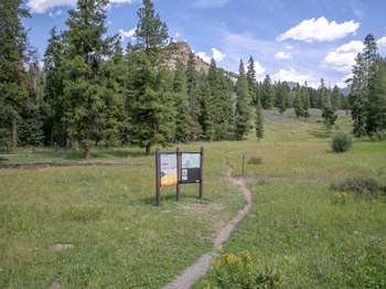 A large trailhead sign stands to the left of the trail the crosses the alpine valley.