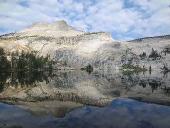 A mountain peak and puffy clouds reflect in a calm, glassy lake.