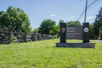 This image is of the Bolivar Heights sign. The sign is brown with a large National Parks Logo. On the left side of the photo there is a brown split rail fence. Trees line the fence and the ground is grassy.