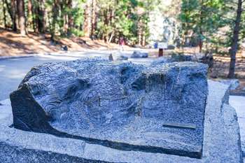 A bronze tactile relief model sits on a granite rock pedestal along a paved trail leading toward a tall waterfall.
