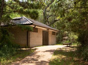 Brown cement structure with a brown roof. Wooden bench on a cement walkway in front of the structure. Green trees surround the structure.