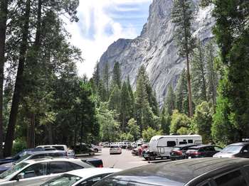 Cars line a parking lot that is surrounded by tall trees. A large granite rock wall rises above the treeline.