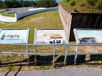 Three metal framed National Park Service signs. The white Spanish Water Battery and brick Fort Barrancas in the background.