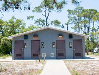 A grey stone building with four brown doors. Four brown handicap and shower signs hang next to each door. A sidewalk on green grass in the foreground. Green trees and blue sky in the background.
