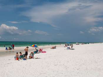 People sitting on towels and under umbrellas on a white sand beach. Turquoise water and blue sky in the background.