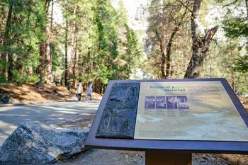 A wayside exhibit with a bronze tactile relief section stands next to a trail that leads toward a tall waterfall.