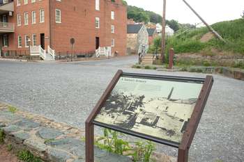 Wayside near a street. To the left of it is a large, red brick building with several white windows. Directly in front of the wayside is the end of Shenandoah Street and the start of Potomac Street.