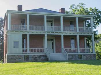 This image shows a large, brick house with white doors, steps and balcony. There are two brick chimneys and white columns in the center and sides on the front of the house. In front of the house is a large yard.