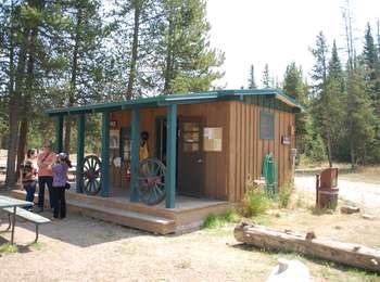 Visitors outside the office of the Colter Bay Corrals. Two wagon wheels decorate the porches. A picnic table and trash bin and nearby.